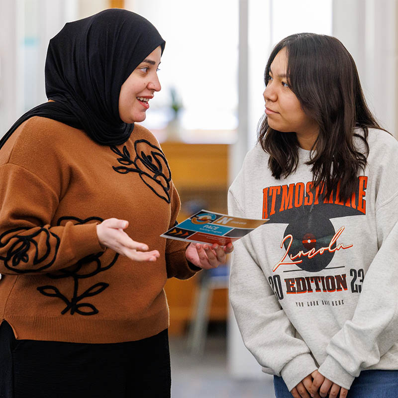 Two people talking indoors, one holds a brochure and gestures; casual clothing visible.