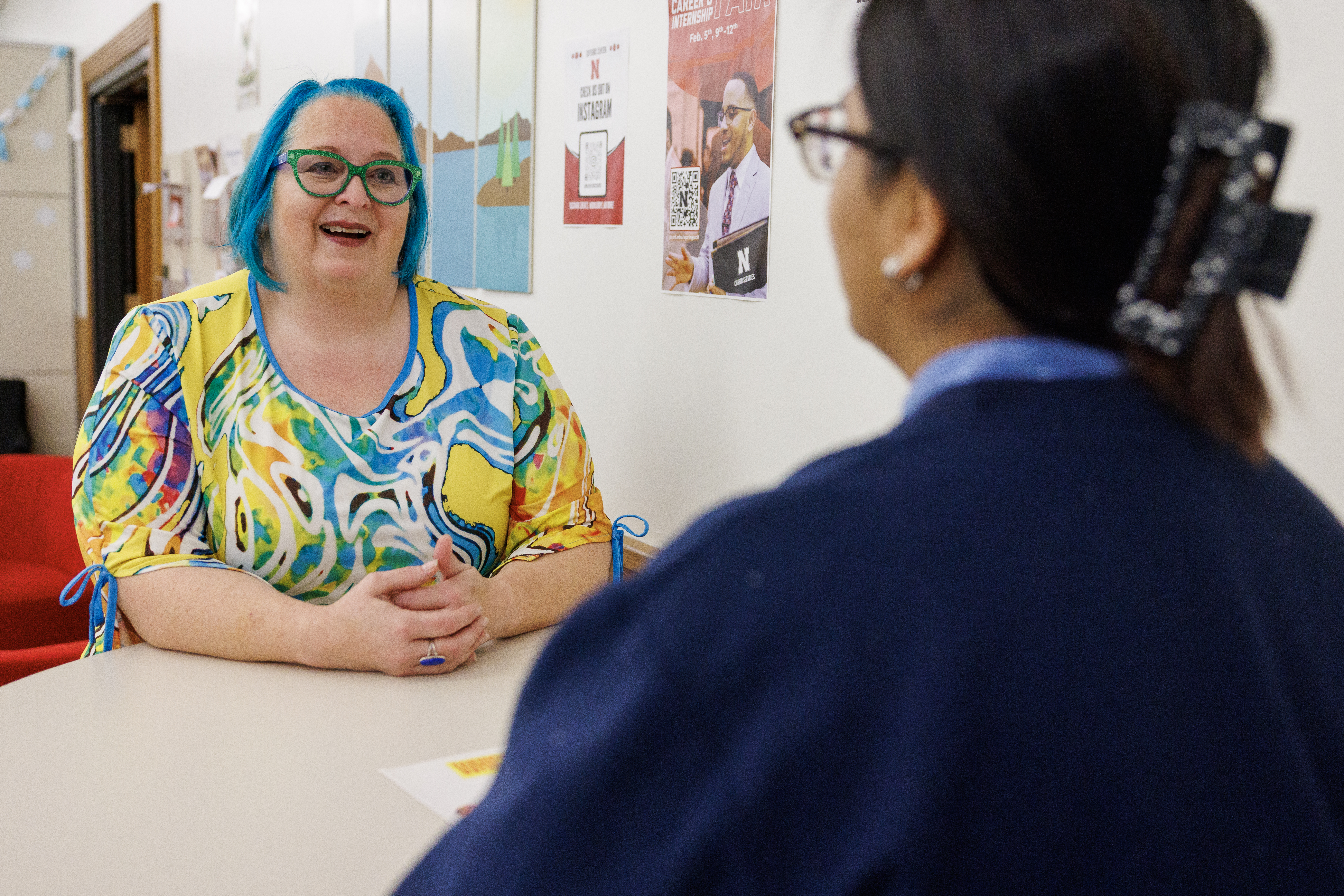 A woman with blue hair and a colorful shirt talking to another person in an office setting.